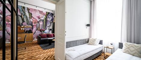 A hotel room with a herringbone floor, a bed with a grey headboard, a bedside table with a wine glass, and a view of a cityscape through the window.
