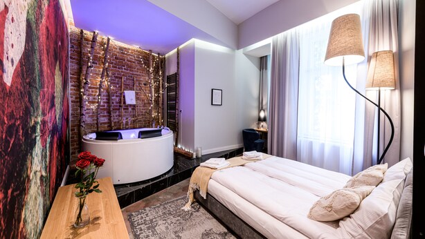 Ein modernes Hotelzimmer mit Ziegelfassade, einem großen Bett, einer Sitzecke mit Tisch und einem Wellnessbereich mit Badewanne.