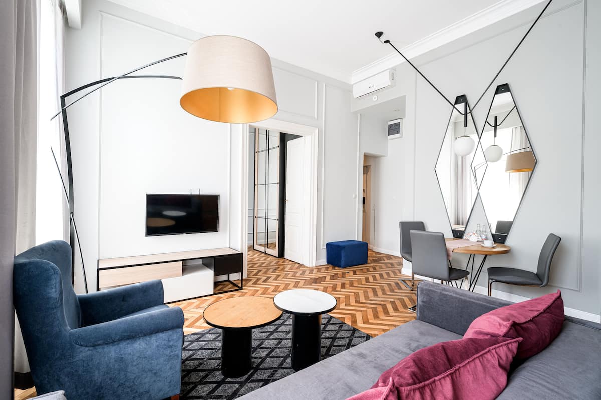 A modern living room with a sofa, armchair, coffee table, and a TV. There is a large mirror on the wall and a dining area in the background.