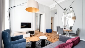 Ein modernes Wohnzimmer mit einer Couch, einem Sessel, einem Couchtisch und einem Fernseher. An der Wand befindet sich ein großer Spiegel und im Hintergrund ein Essbereich.