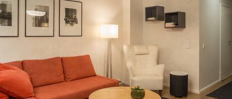 A modern living room with a red sofa, a white armchair, a round wooden coffee table, and a floor lamp. There are framed pictures on the wall and a small bookshelf.