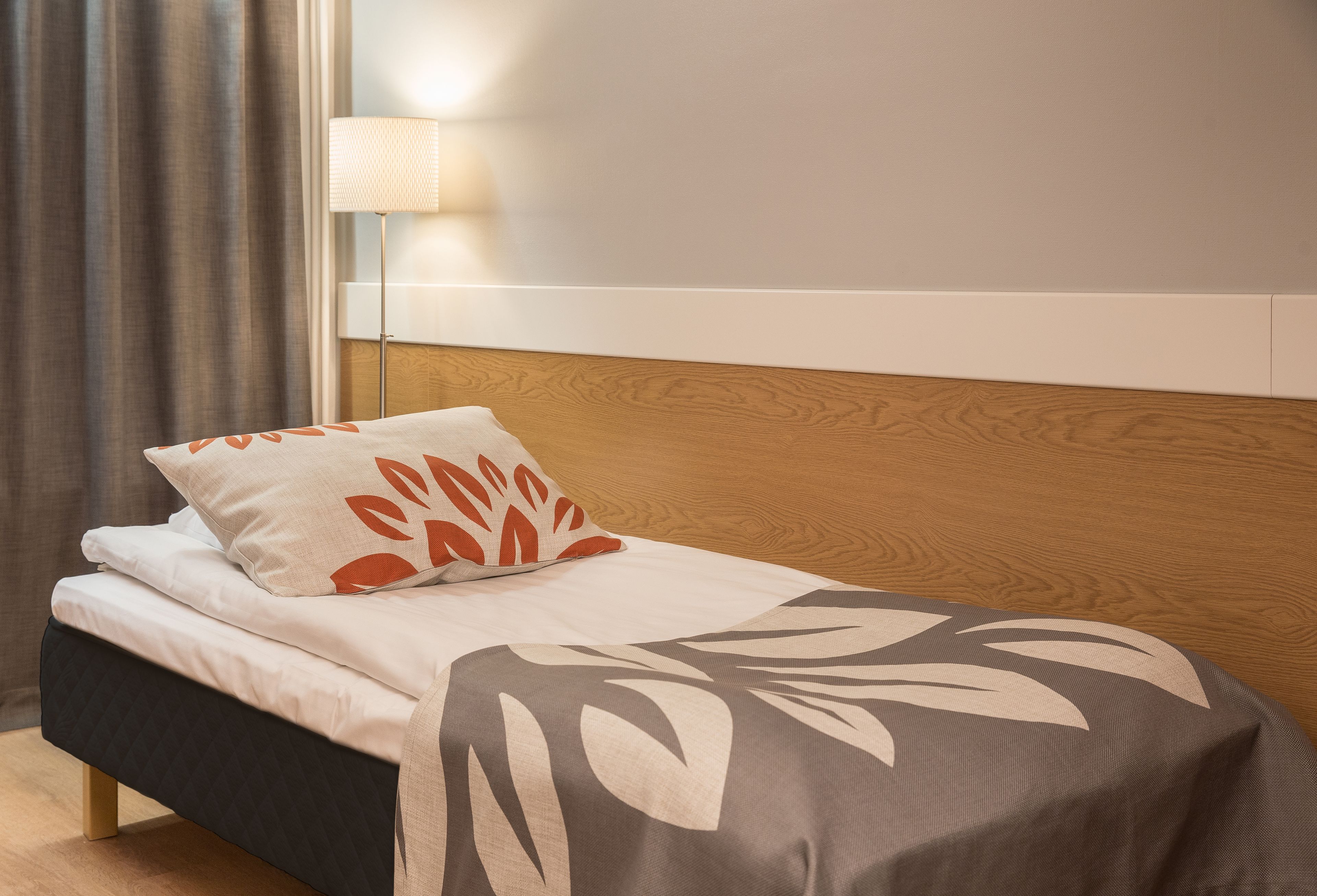 Una cama bien hecha con un edredón estampado, un almohadón con diseño de hojas y una lámpara de noche.