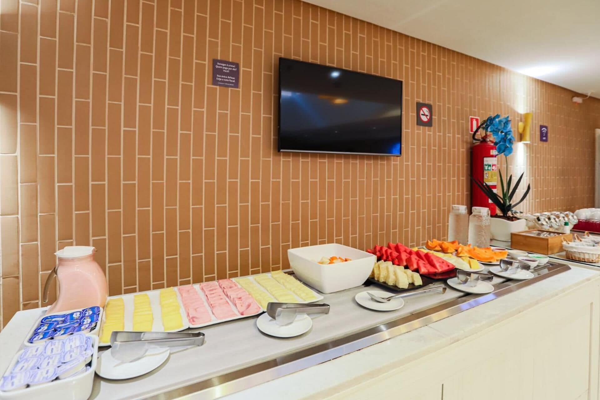 A buffet setup with various food items and utensils, a television mounted on a brick wall, and a fire extinguisher.