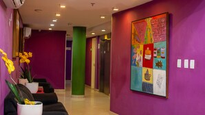 A brightly colored hallway with purple walls, a green pillar, and a colorful artwork on the wall. There are potted plants and a black couch in the foreground. - Rede Andrade Docas (Belém)