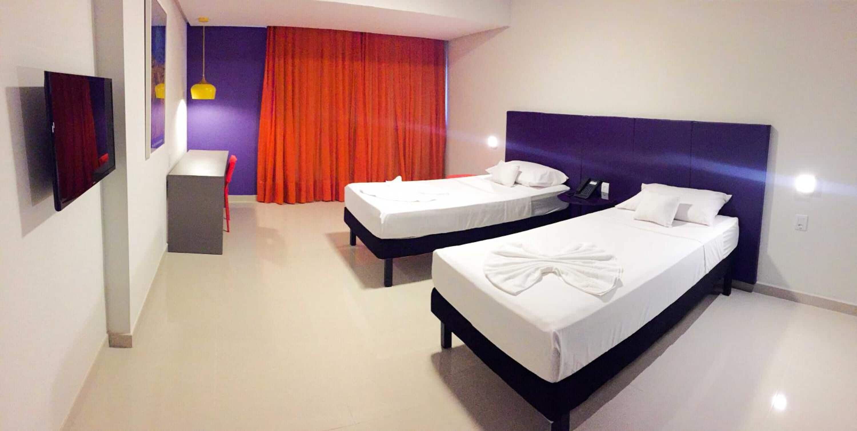 A hotel room with two single beds, a desk, a chair, a TV, and a red curtain.