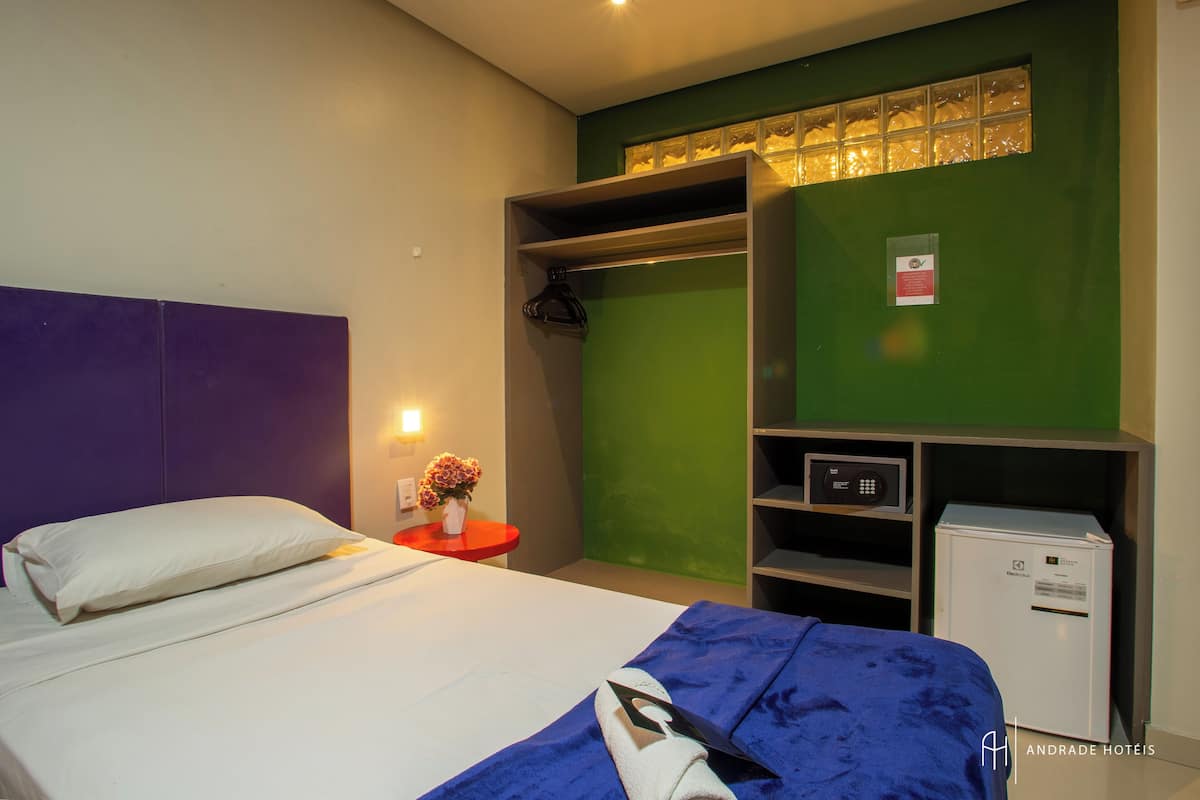 A hotel room with a bed, a red side table with a vase of flowers, a green wall, a wardrobe, and a minibar.