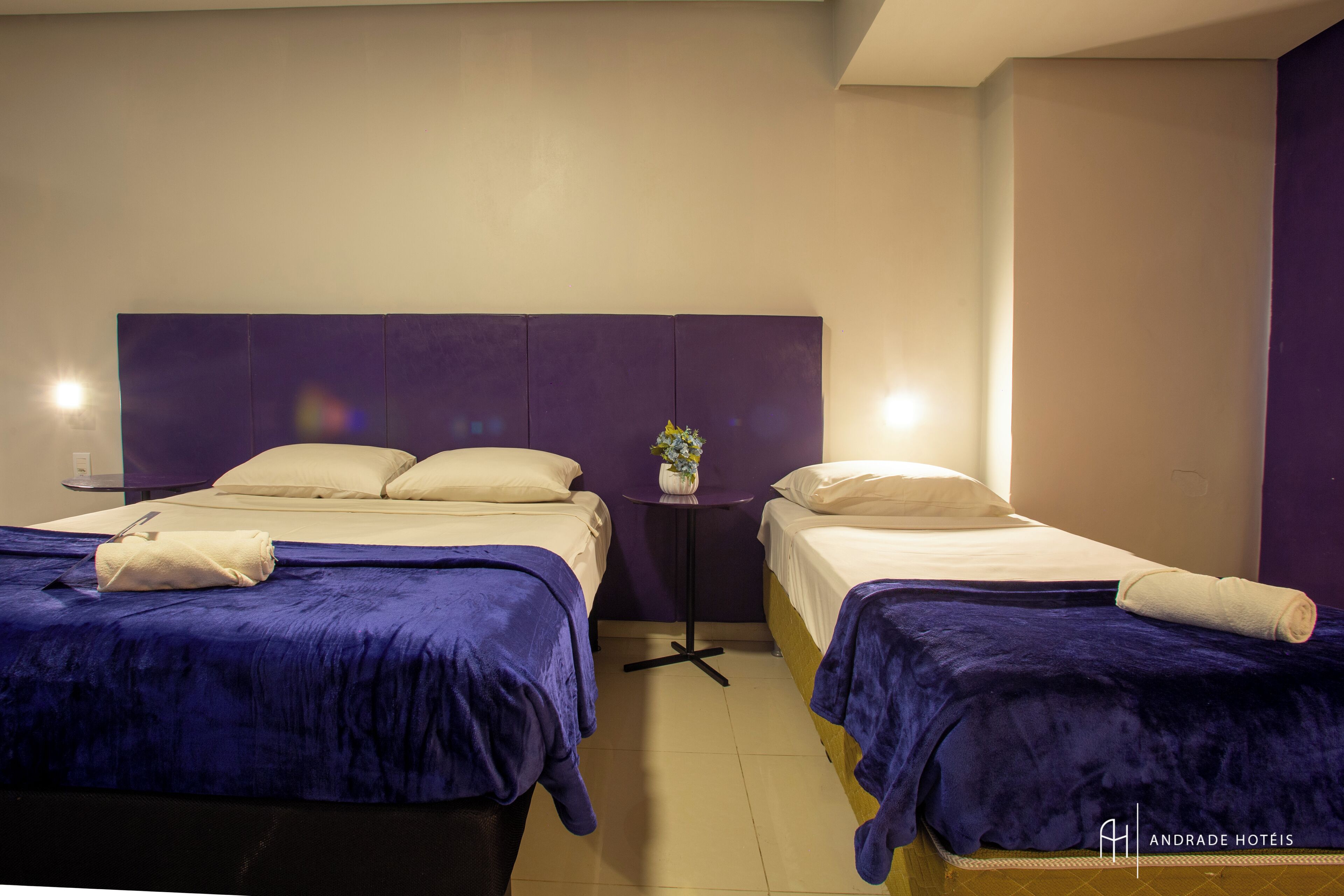 A hotel room with two beds, each with a blue bedspread and white pillows. There is a small table with a vase of flowers between the beds.