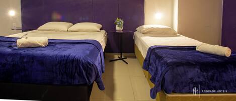 A hotel room with two beds, each with a blue bedspread and white pillows. There is a small table with a vase of flowers between the beds.