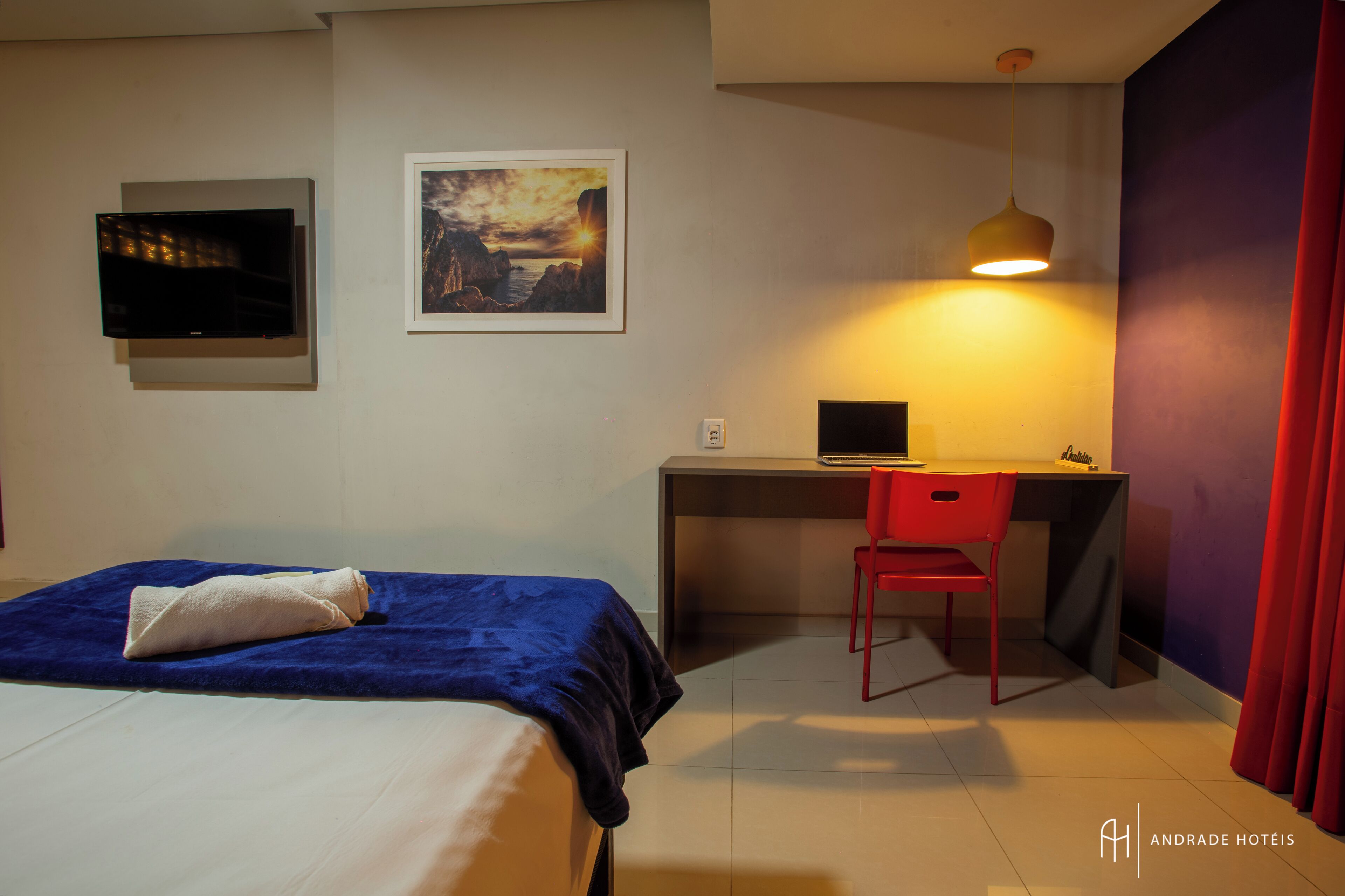 Une chambre d’hôtel avec un lit, un bureau avec un ordinateur, une chaise, une télévision et un tableau au mur.