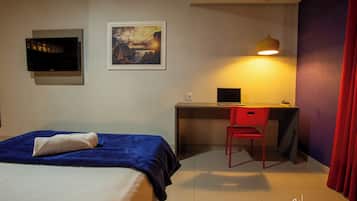 A hotel room with a bed, a desk with a computer, a chair, a TV, and a painting on the wall.