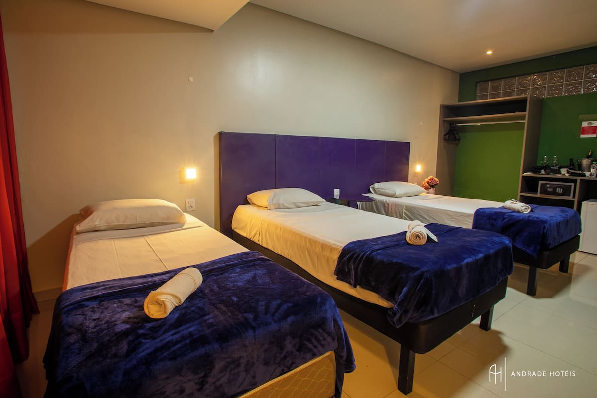 A hotel room with three beds, each with a blue blanket and a rolled towel. There is a small desk with a computer and a green wall with a decorative panel.