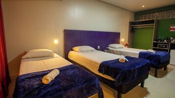 Habitación de hotel con tres camas, cada una con una manta azul y una toalla enrollada. Hay un escritorio pequeño con una computadora y una pared verde con un panel decorativo.