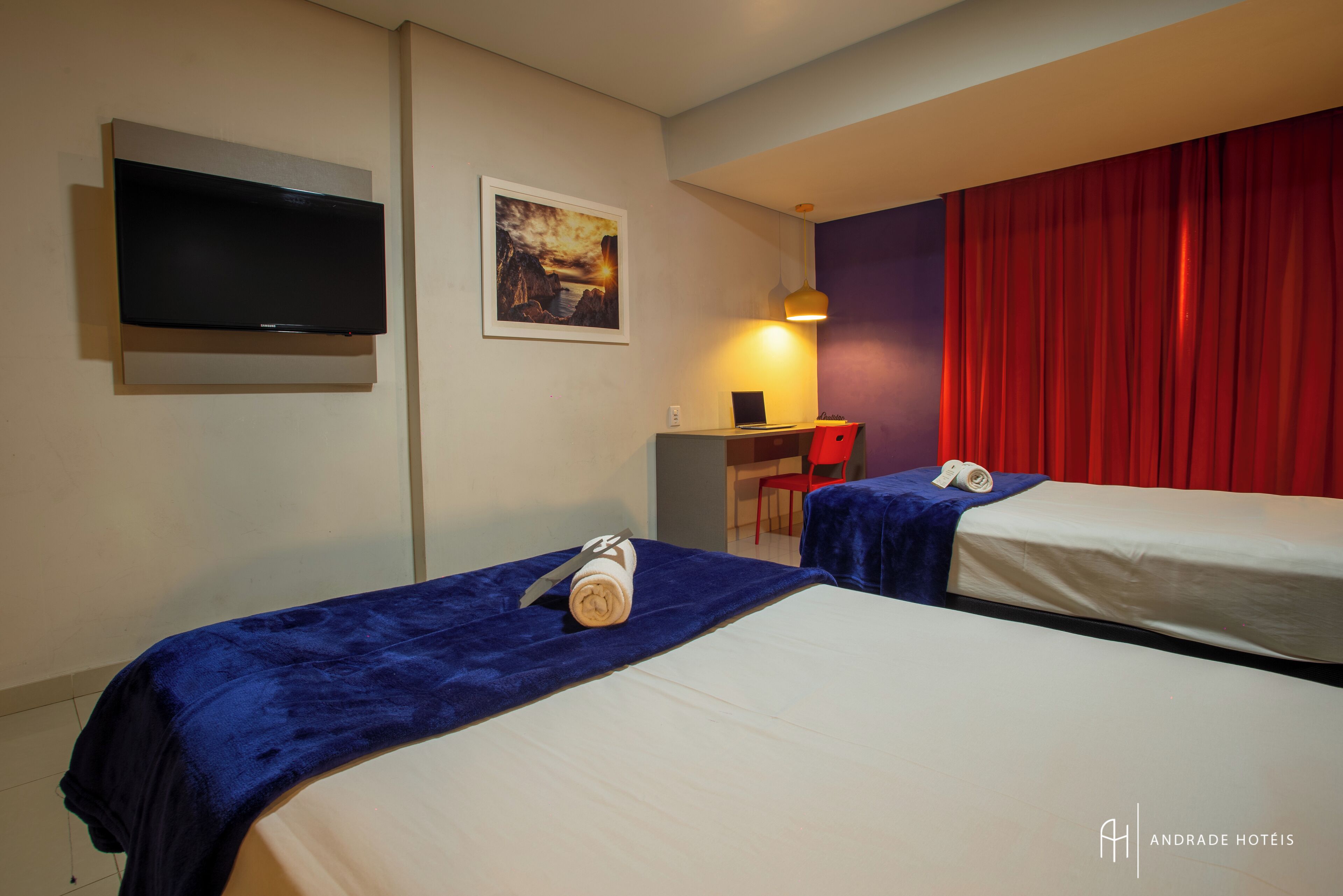 Ein Hotelzimmer mit zwei Betten, einem Schreibtisch, einem Stuhl, einem Fernseher und einem roten Vorhang.