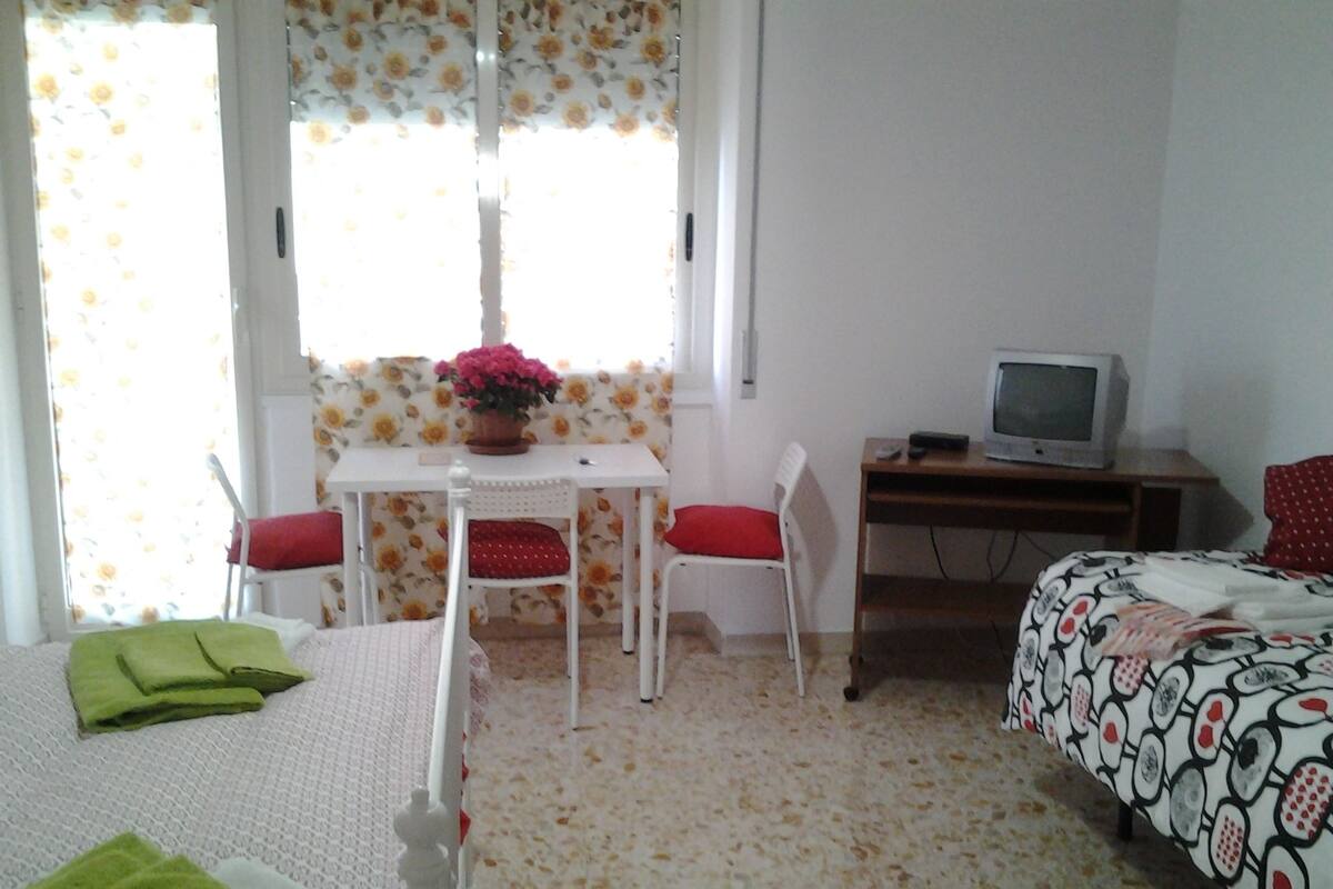 A room with a bed, a desk with a television, a chair, and a window with floral curtains.