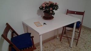 A small dining area with a white table, two wooden chairs, a blue cushion on one chair, and a potted plant with flowers. - I Colori di Salvator Rosa (Naples)