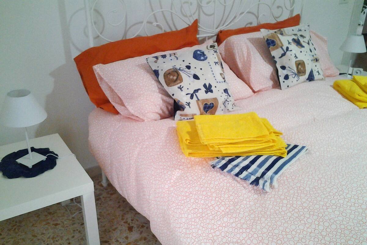 A neatly made bed with pink bedding, orange pillows, and a blue and white striped pillow. There is a white nightstand with a lamp and a folded yellow towel.