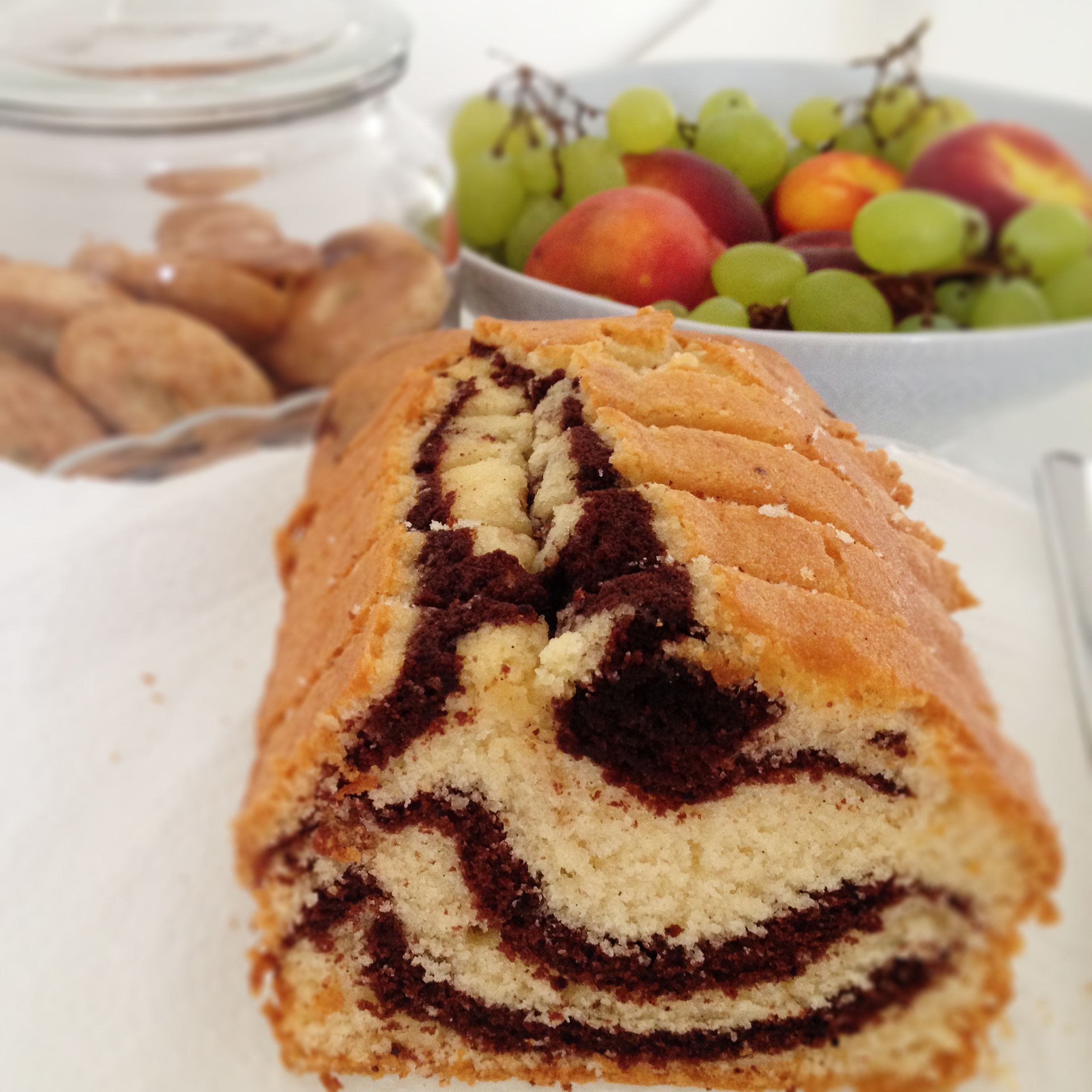 A marble cake with a visible swirl pattern, a bowl of fruit, and a jar of cookies in the background.