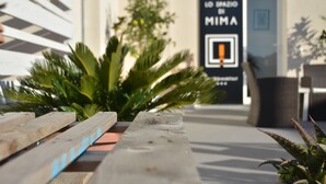 A wooden bench with plants in the foreground, a door with a sign reading "LO SPAZIO MIIMA," and a potted plant to the right. - Lo spazio di Mima (Noto)