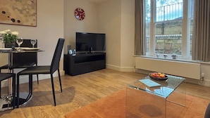 A modern living room with a dining area, a flat-screen TV, a large window with curtains, and a decorative clock. - Your Space Apartments - Jubilee House (Cambridge)