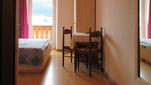 A bedroom with a bed, a wooden chair, a small table, a TV mounted on the wall, and a balcony with a view. - Albergo Lucia (Tesero)