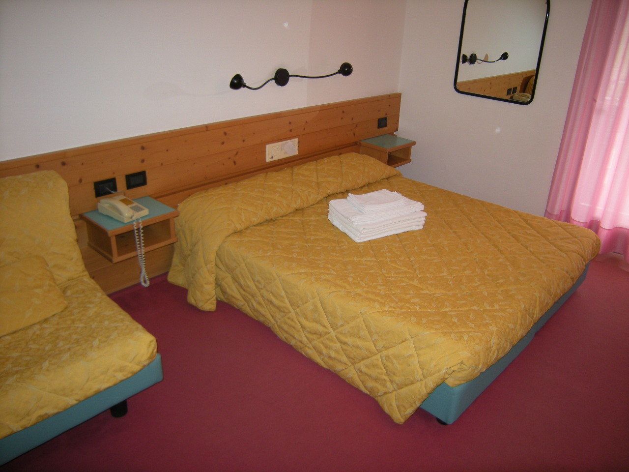 A hotel room with two beds, a wooden headboard, a bedside table with a lamp, and a mirror on the wall.