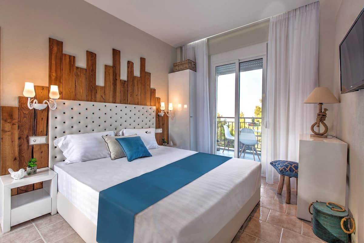 A hotel room with a large bed, wooden headboard, white nightstands, a blue accent pillow, and a balcony with a chair.