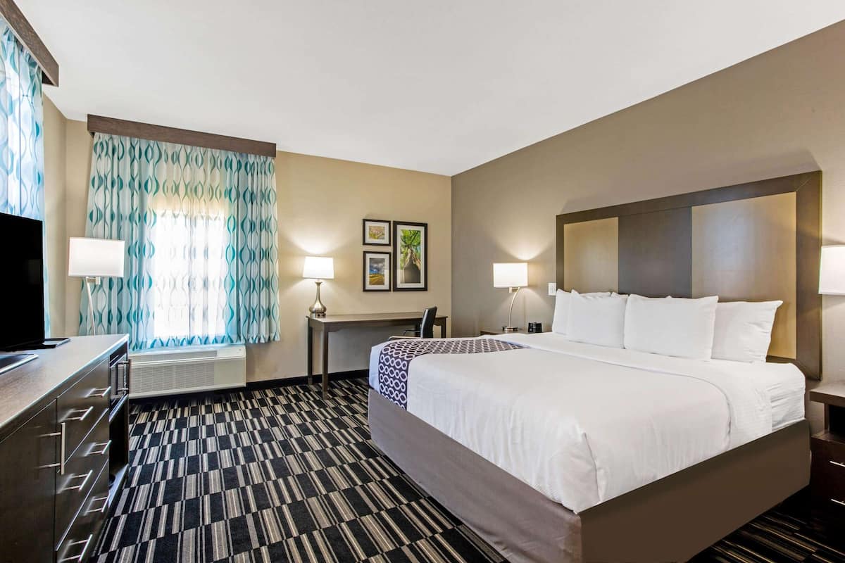 Ein Hotelzimmer mit einem großen Bett, einem Schreibtisch mit Stuhl, einem Flachbildfernseher und einem Fenster mit gemusterten Vorhängen.