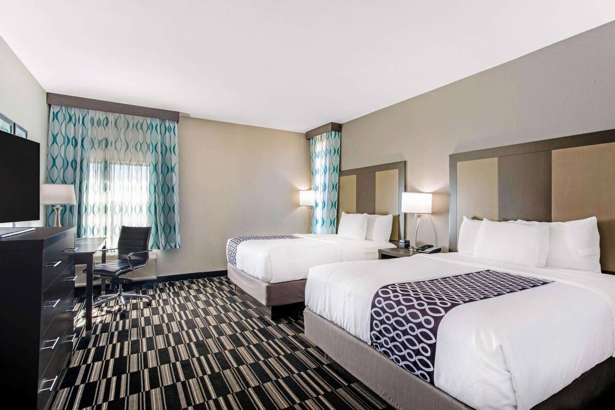 Ein Hotelzimmer mit zwei Betten, einem Schreibtisch mit Stuhl, einem Fernseher und einem gemusterten Teppich.