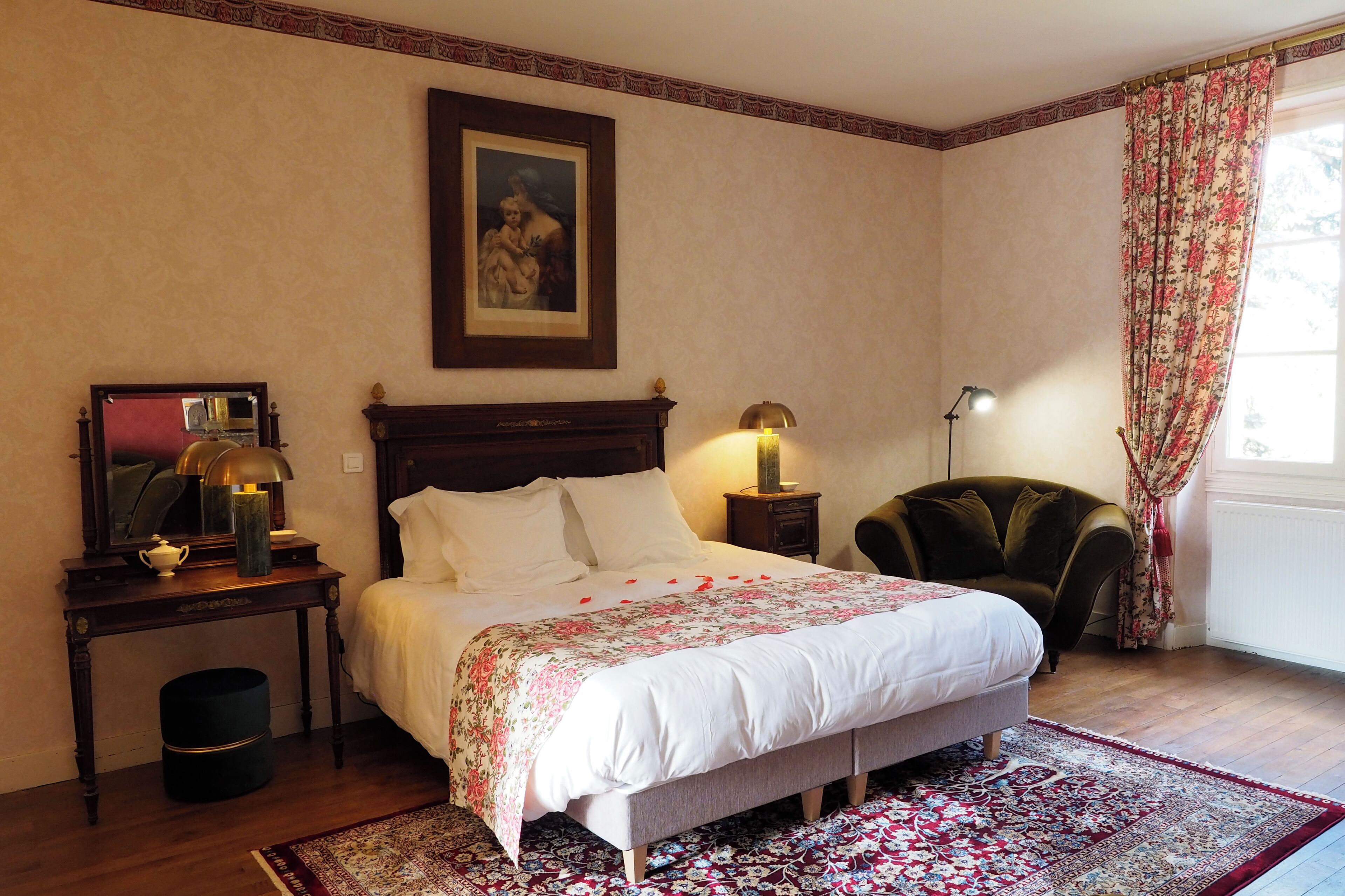 Un dormitorio con cama, mesitas de noche, lámpara, silla, alfombra y ventana con cortinas.
