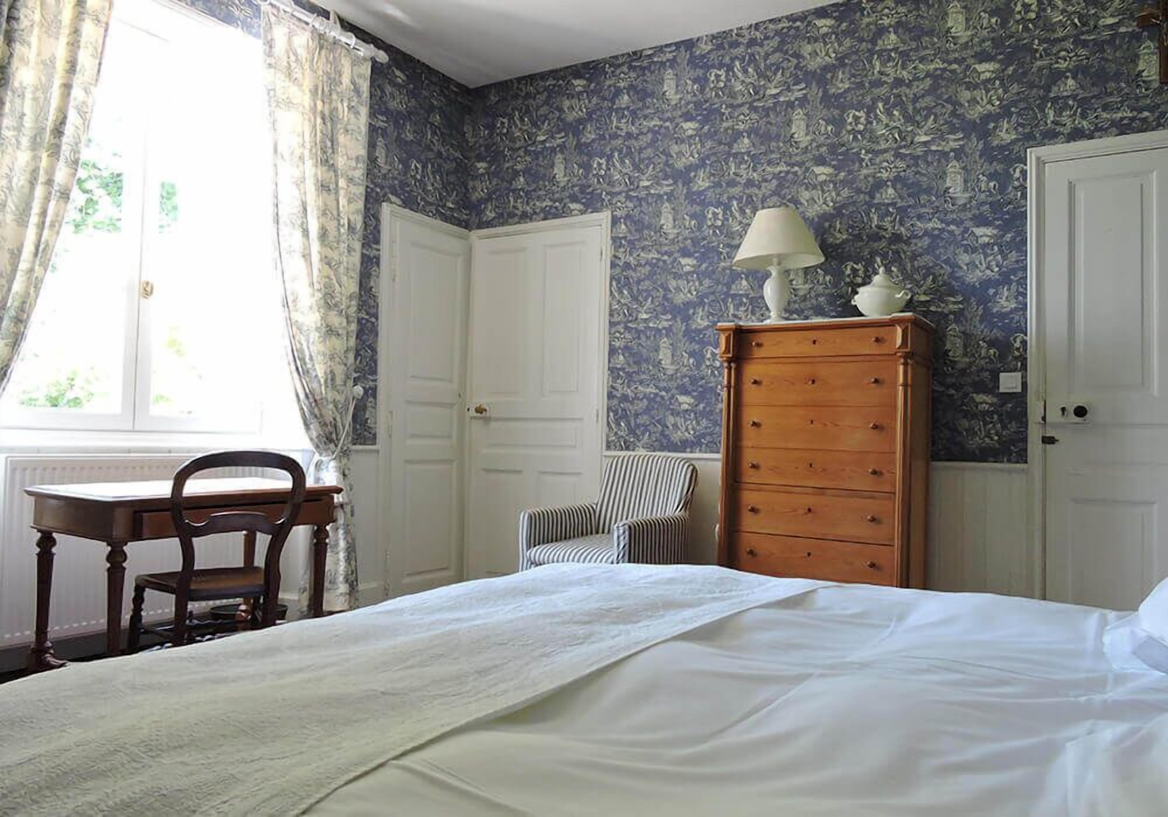 Ein Schlafzimmer mit einem Bett, einem Holzkleiderschrank, einem Stuhl, einem Schreibtisch und einem Fenster mit Vorhängen.