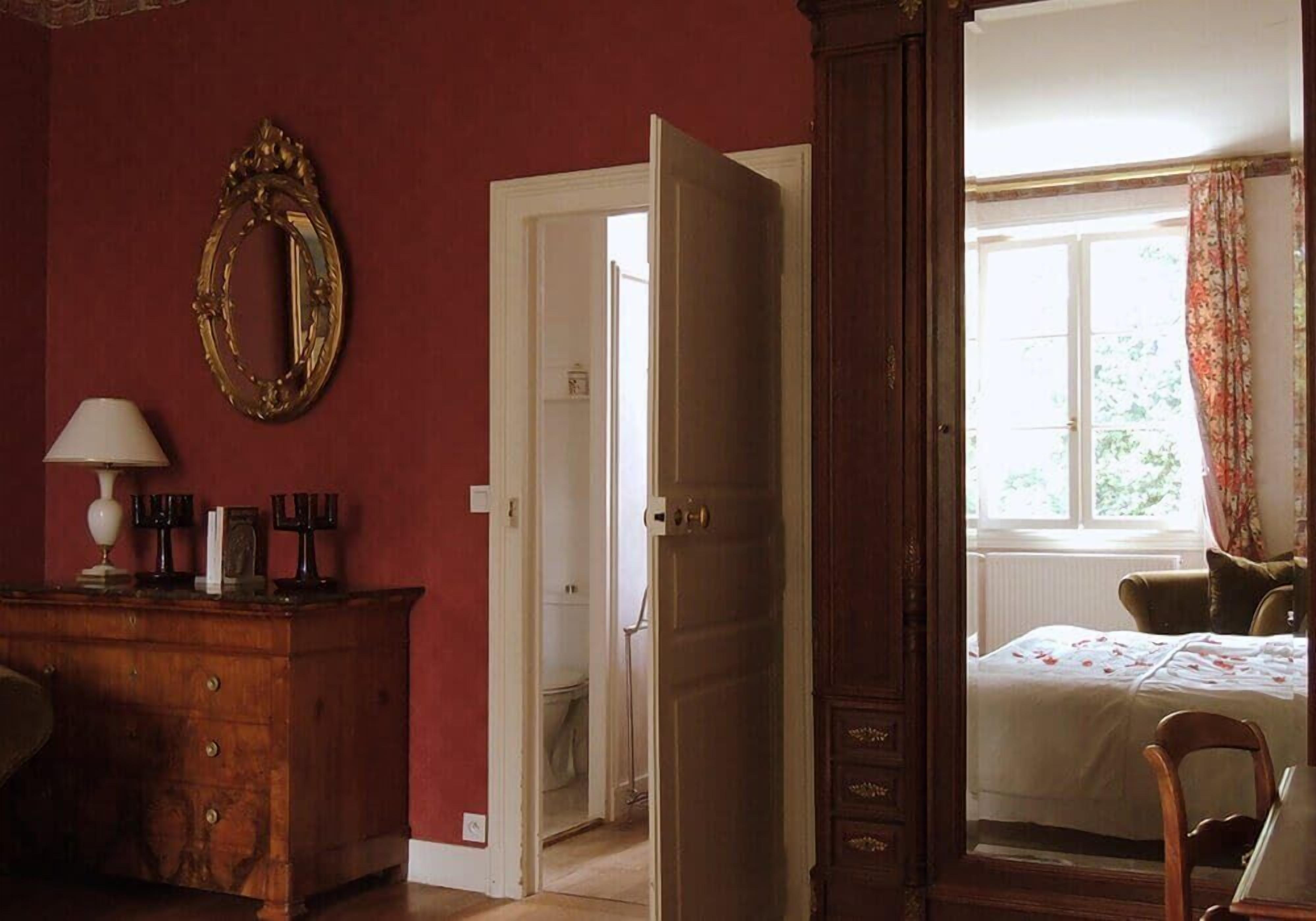 Una habitación con un aparador de madera, un espejo, una cama con colcha estampada y una ventana con cortinas.