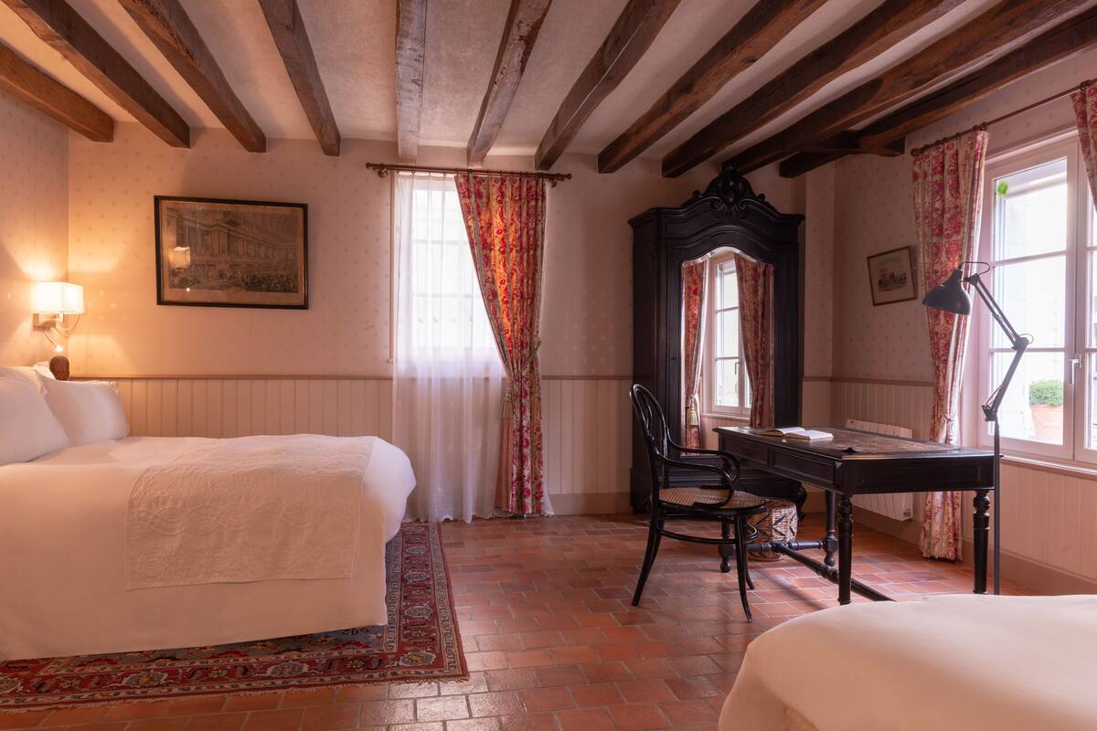 Une pièce comprenant un lit, un bureau et une chaise. Il y a une fenêtre avec des rideaux, une lampe et un tableau encadré au mur.