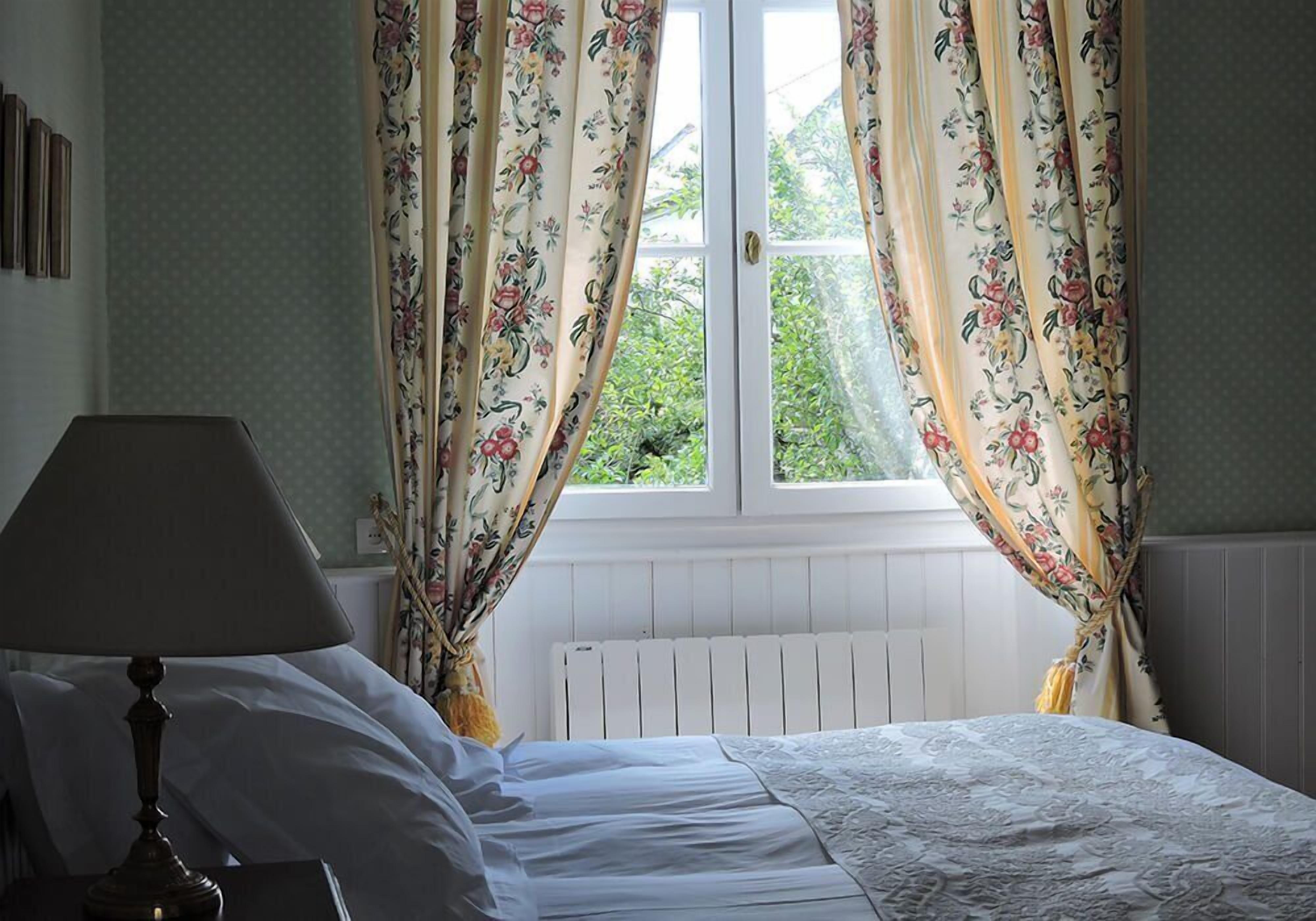 A bedroom with a bed, a window with floral curtains, a radiator, and a lamp.