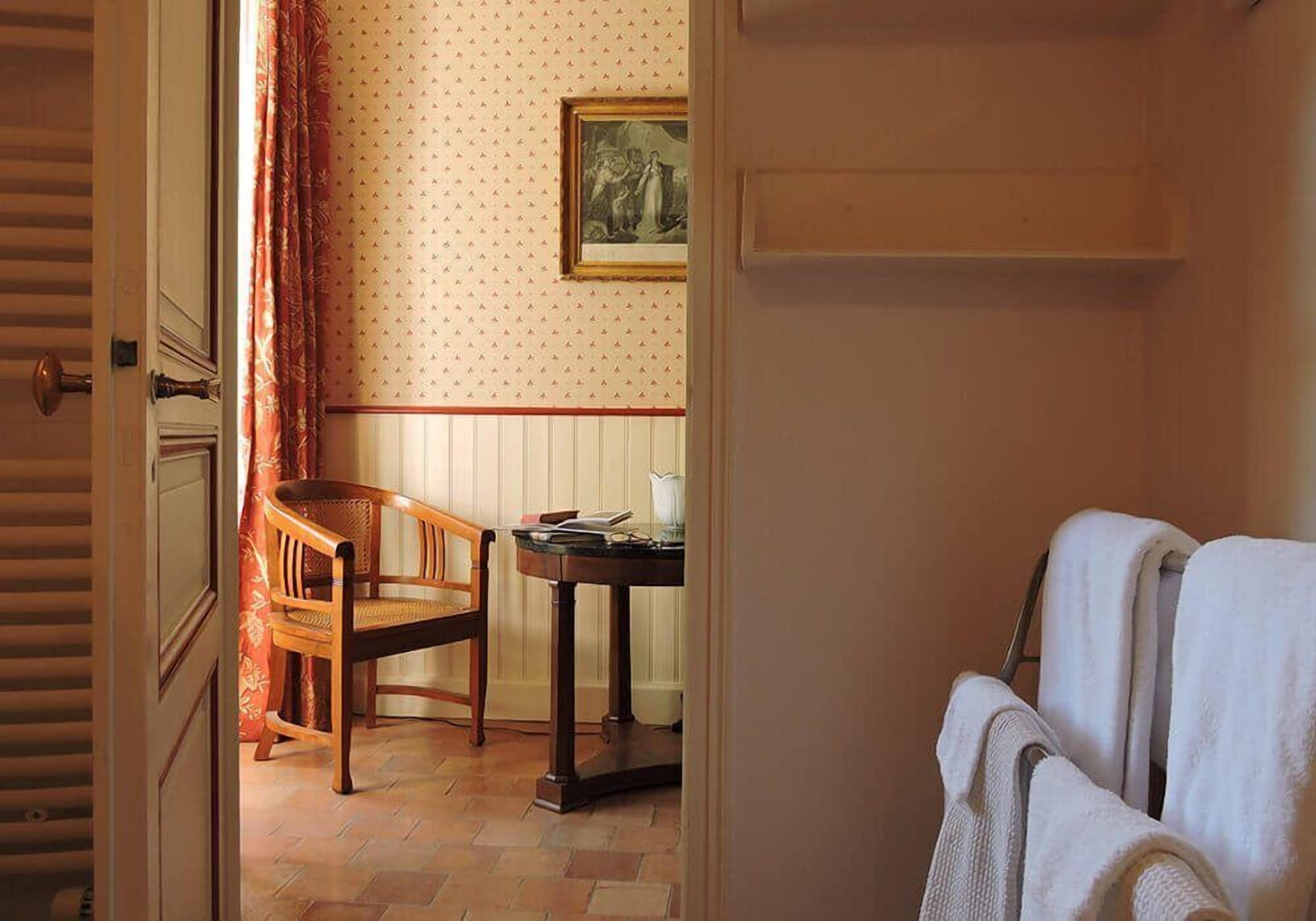 Ein Zimmer mit einem Holzsessel, einem kleinen Tisch mit einem Buch und einem weißen Handtuch an einem Wäscheständer.
