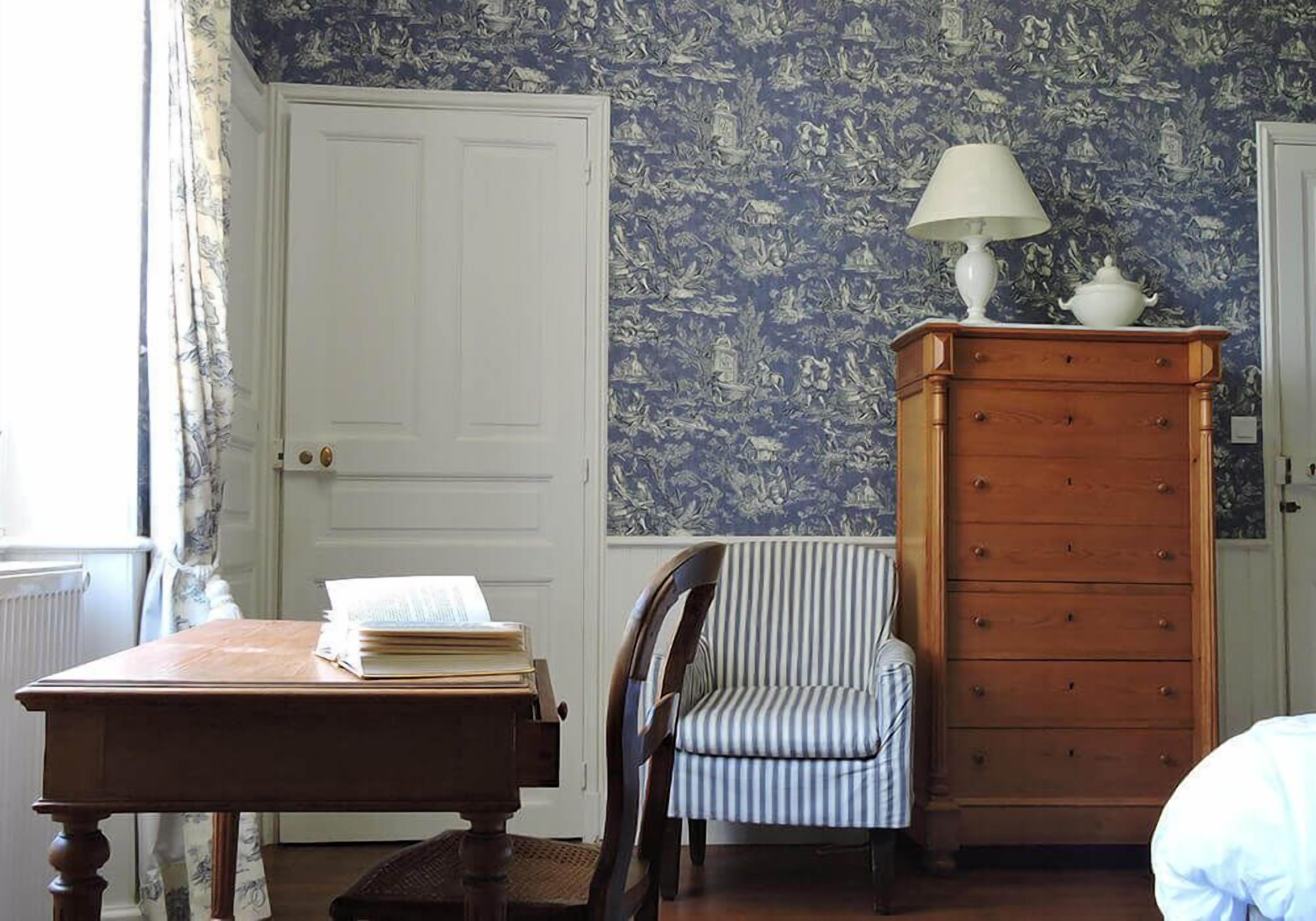 Una habitación con un escritorio de madera, una silla a rayas, un aparador con una lámpara y un papel tapiz estampado.