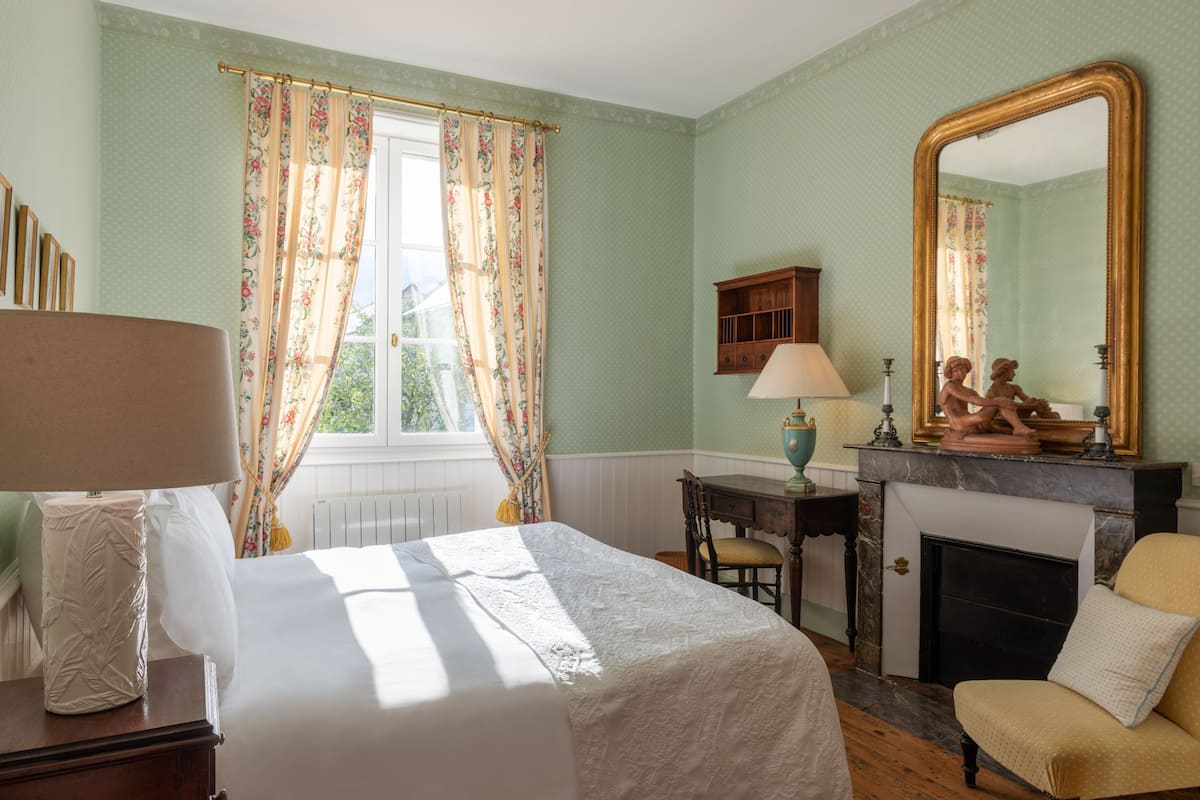 Une pièce comprenant un lit, un bureau, une chaise, une cheminée et un grand miroir.