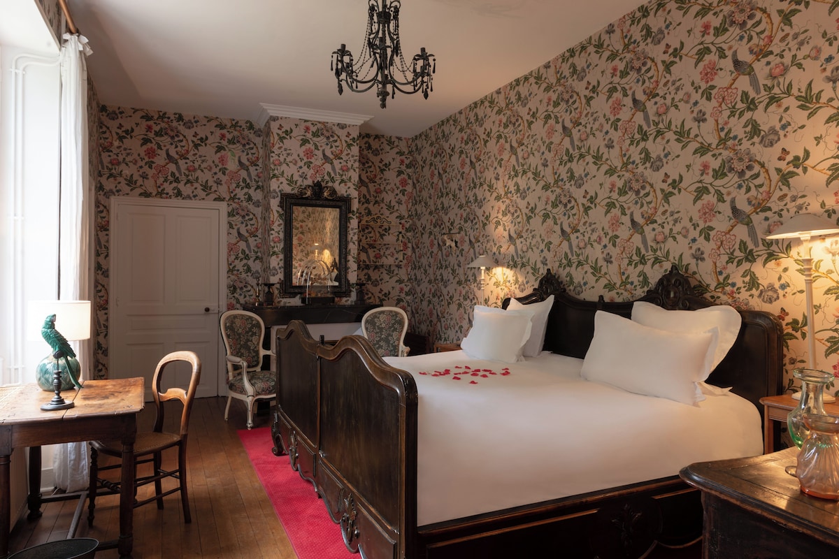 Ein Schlafzimmer mit blumigem Wandpapier, einem hölzernen Bett, einer Chandelierleuchtre, einem Schreibtisch mit Stuhl und einem Spiegel.