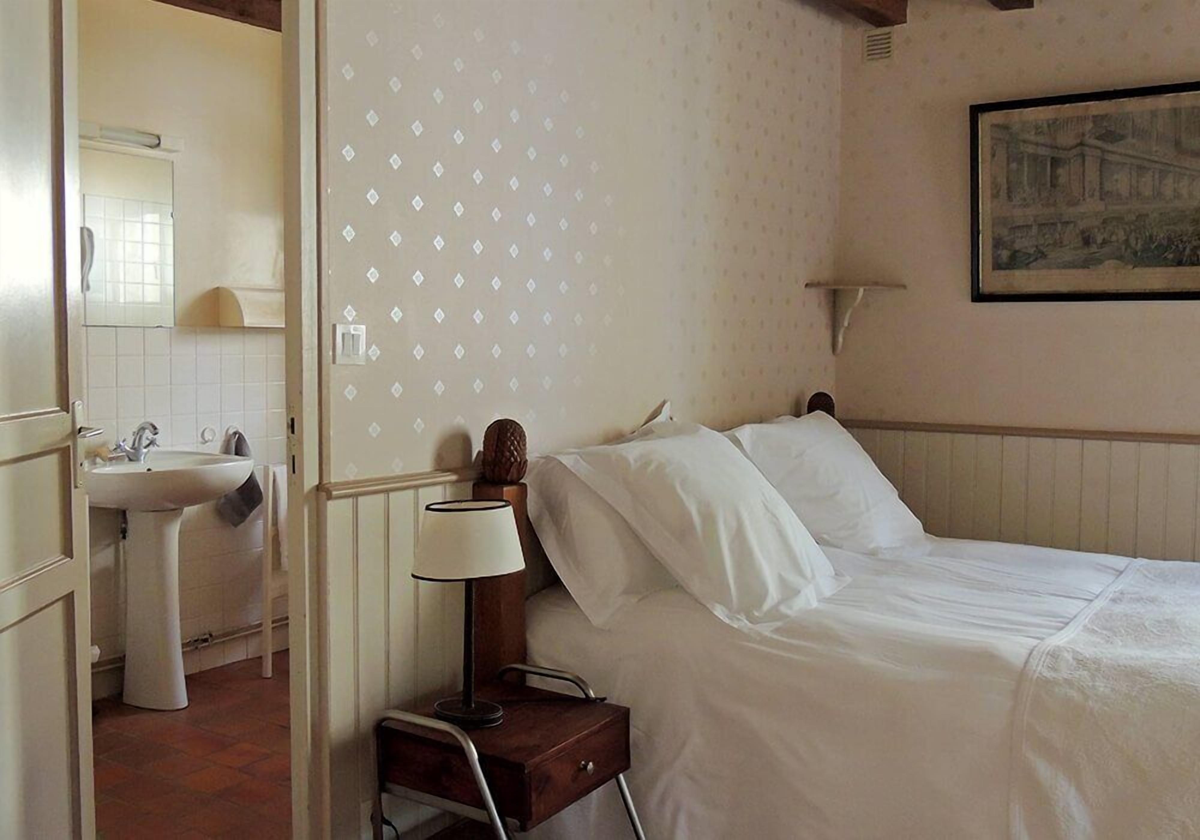 Un dormitorio con cama, mesita de noche con lámpara, cuadro en la pared y baño con lavabo y espejo.