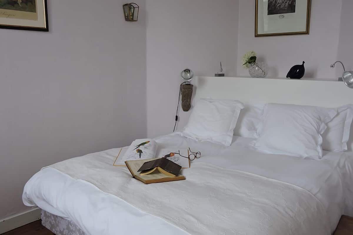 Un lit bien fait, avec du linge de lit blanc, un livre et des lunettes posés dessus. On y voit une lampe, un vase avec des fleurs et un tableau encadré accroché au mur.