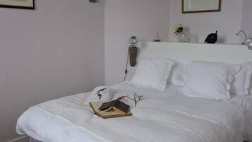 Una cama bien hecha con sábanas blancas, un libro y unos lentes encima. Hay una lámpara, un florero con flores y un cuadro en la pared.