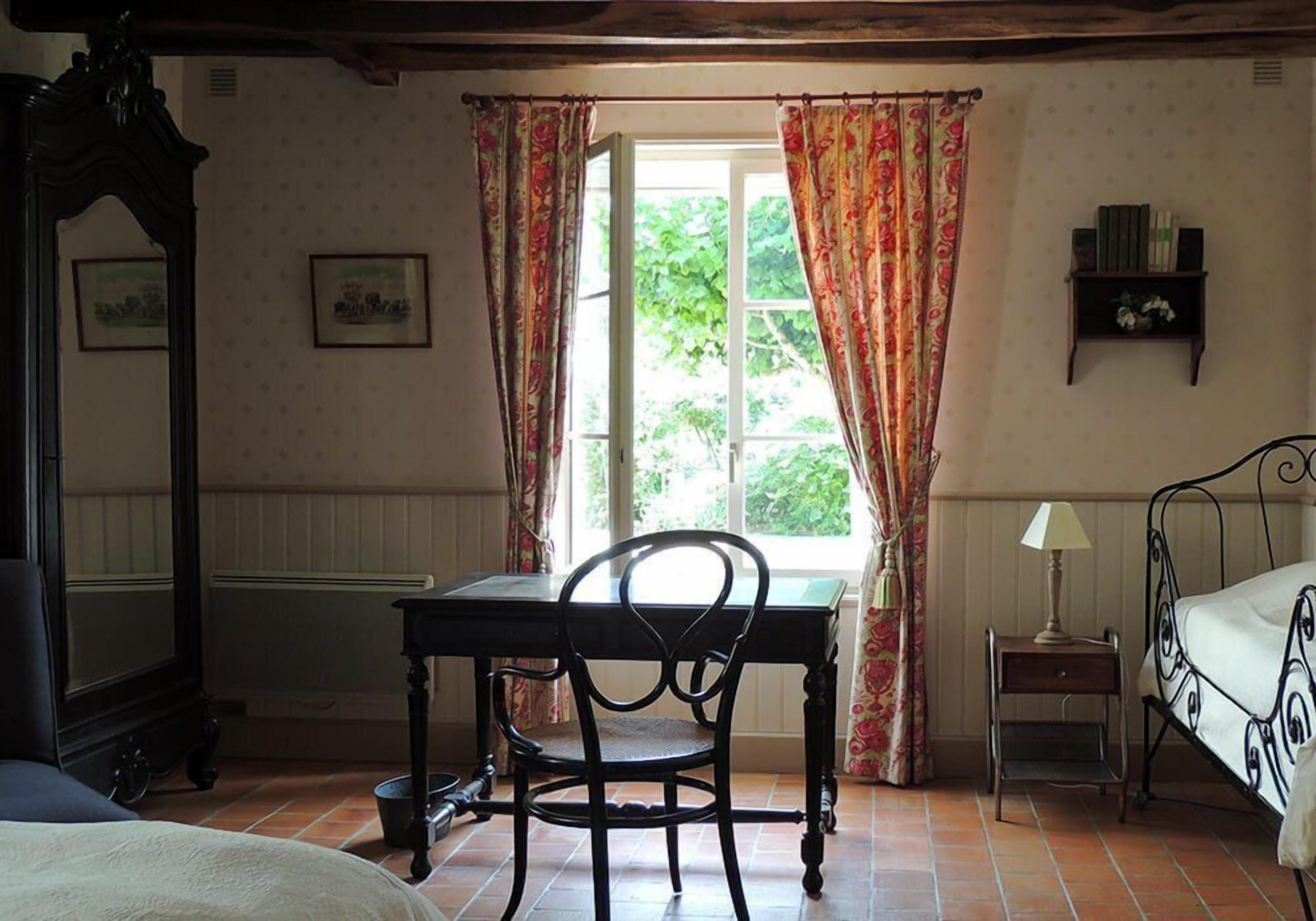 A room with a bed, a desk, and a chair. There is a window with curtains, a radiator, and a shelf with books.