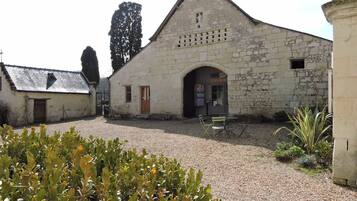 Ein Steinhaus mit einem zentralen Bogenportal, einem kleinen Fenster und einer Tür. Vor dem Haus führt ein Kiesweg, und rechts ist eine Bank zu sehen.