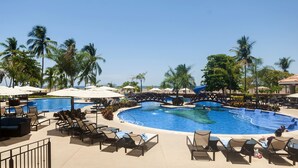 Un espace piscine aménagé dans un complexe hôtelier, avec de nombreux chaises longues et parasols, le tout entouré de palmiers.