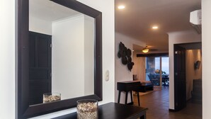 Un couloir moderne avec un grand miroir, un vase en verre posé sur une console, et un coin repas à l’arrière-plan.