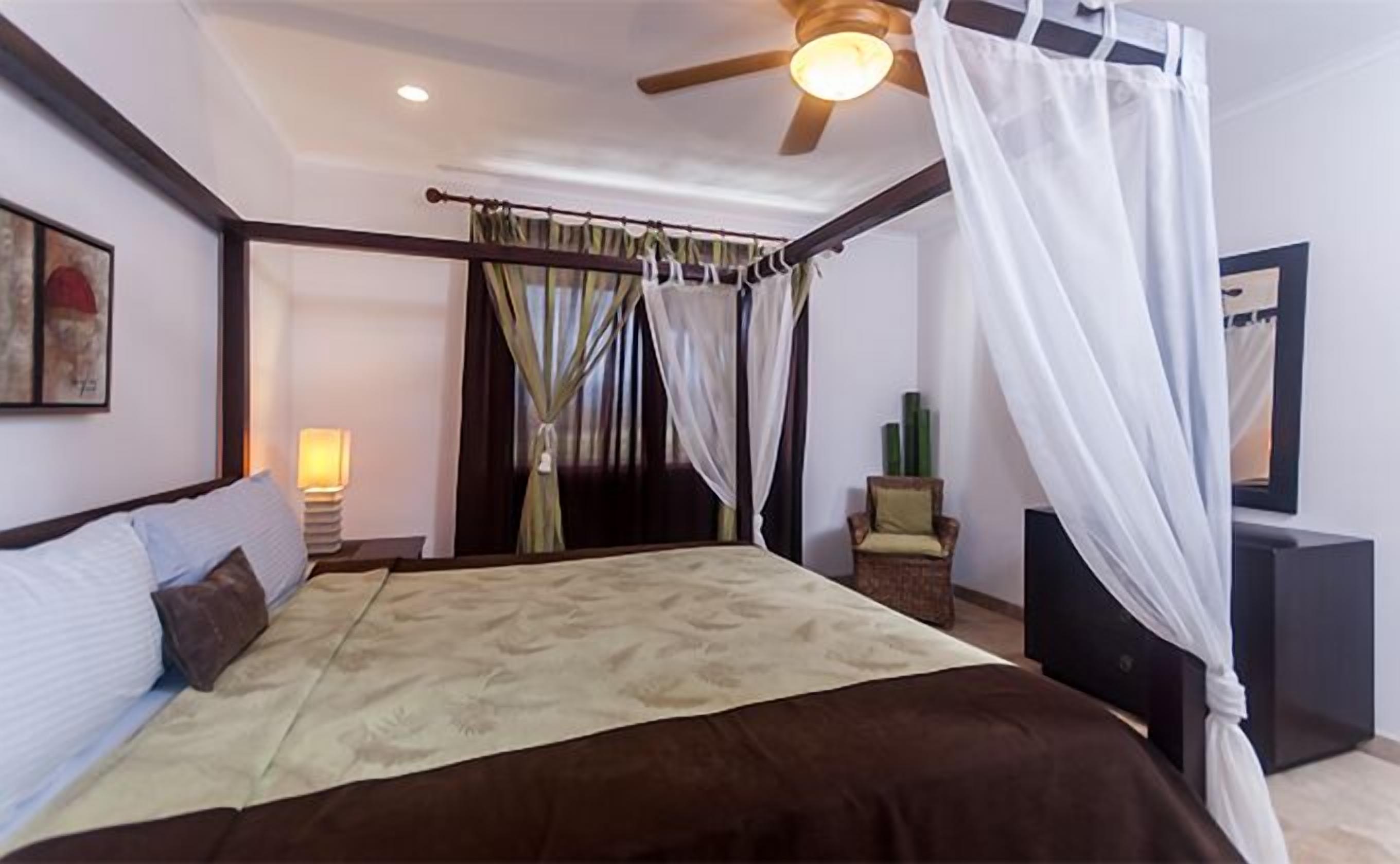 A bedroom with a four-poster bed, a ceiling fan, a bedside lamp, a chair, and a mirror.
