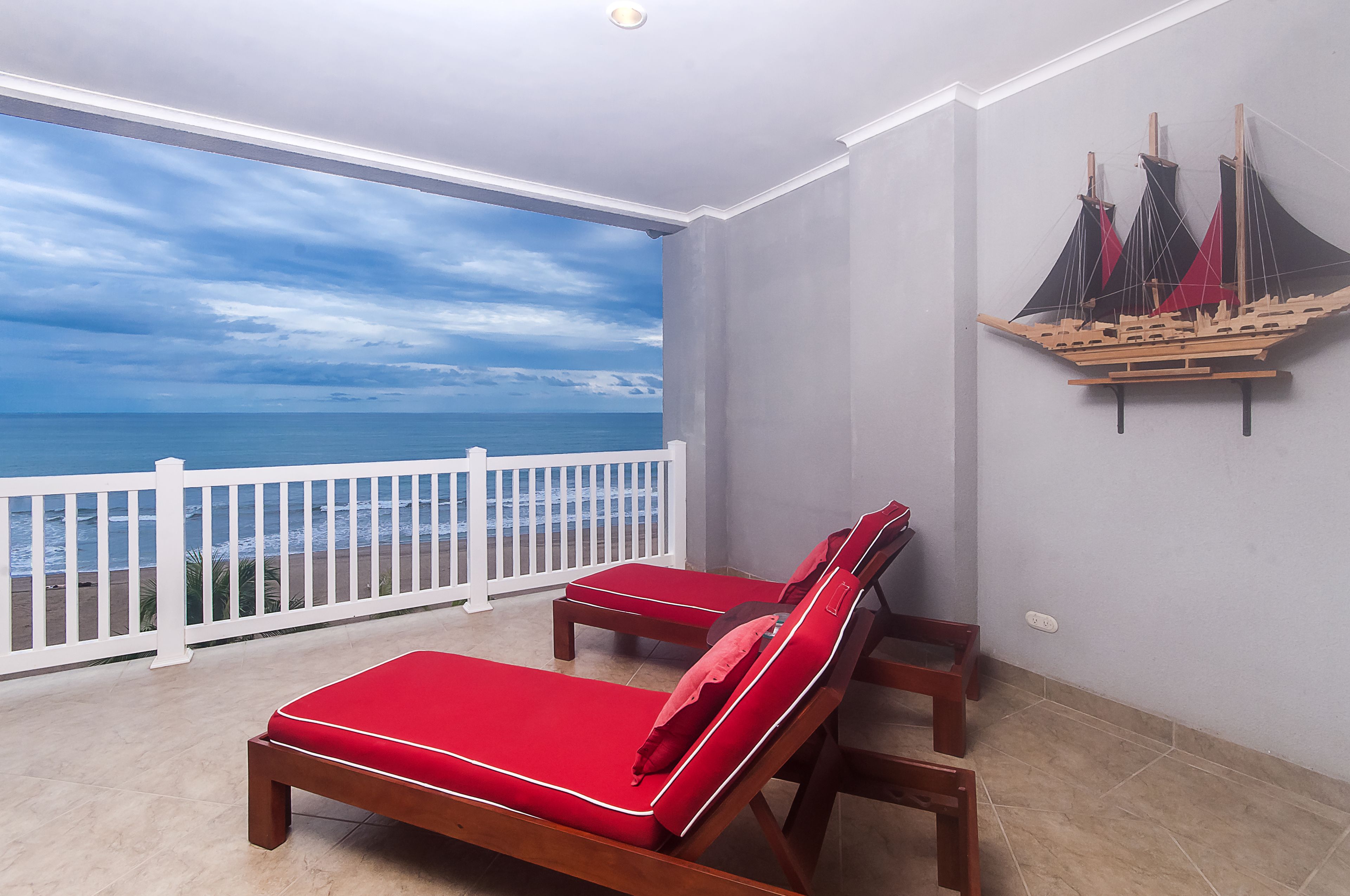 A balcony with two red cushioned lounge chairs, a white railing, and a view of the ocean.