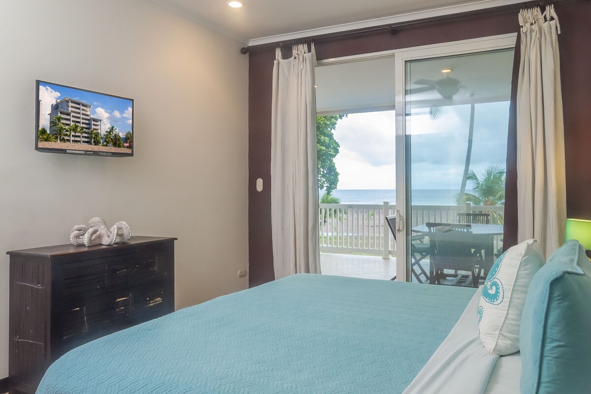 A hotel room with a bed, a dresser, a TV on the wall, and a balcony with a view of the ocean.