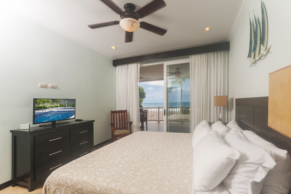 A hotel room with a bed, a TV on a dresser, a ceiling fan, a balcony with a view of the ocean, and a painting on the wall.