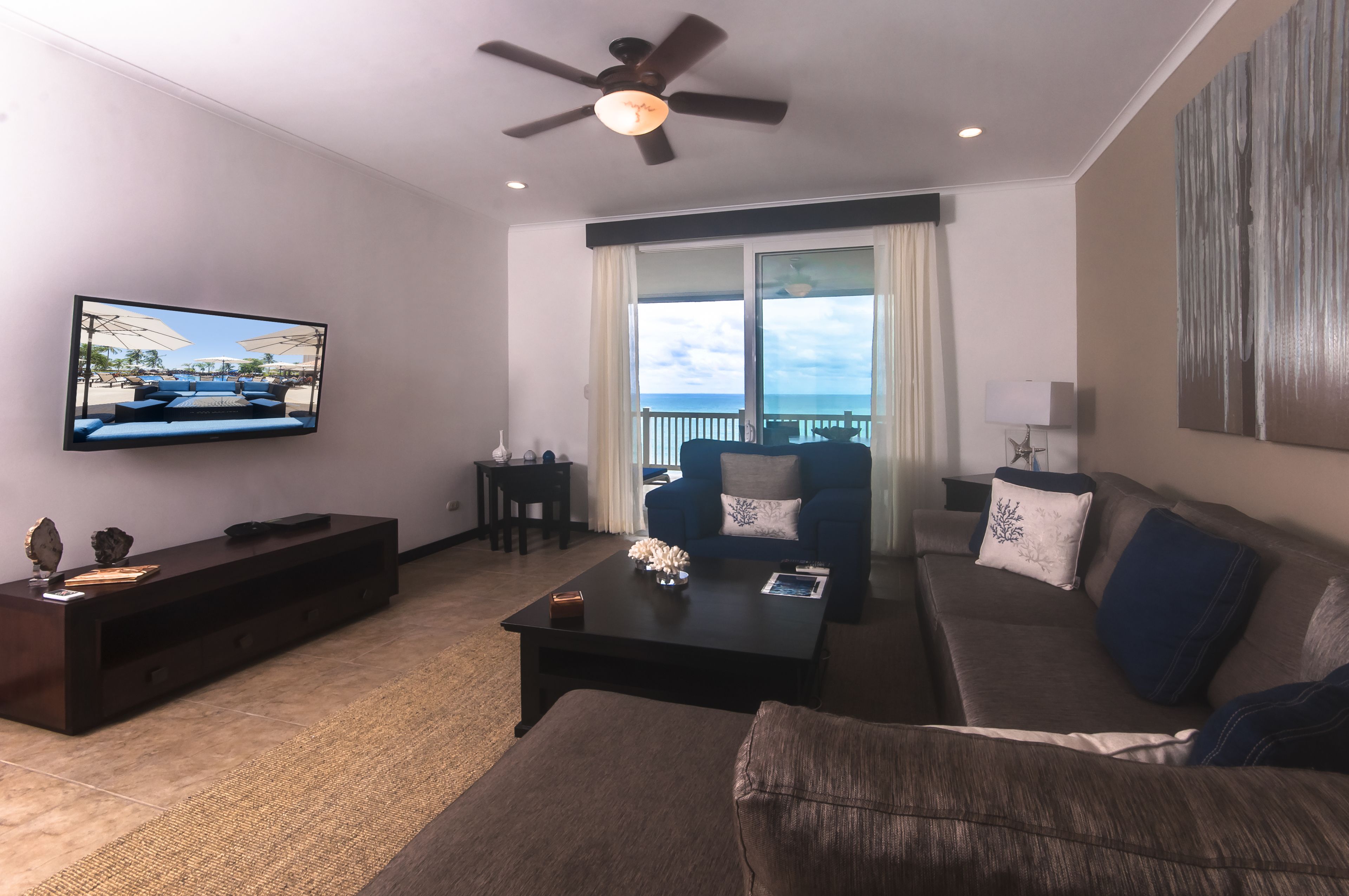 A modern living room with a ceiling fan, a flat-screen TV, a dark wood entertainment unit, a coffee table, a sofa, and a view of the outdoors through large windows.