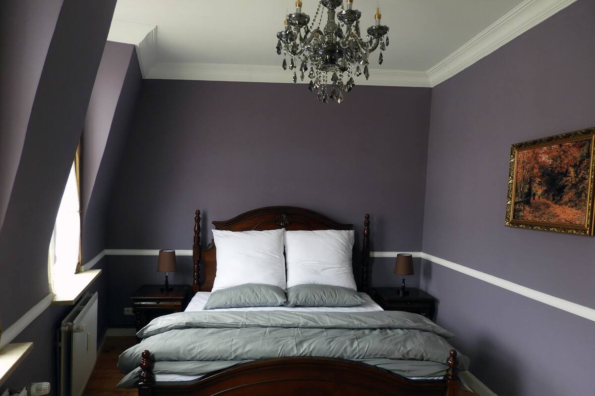 A bedroom with a large bed, two bedside tables, a chandelier, and a painting on the wall.
